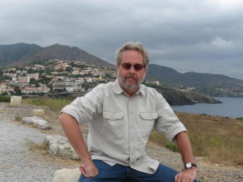 Courtesy image of Larry Ellis, sitting on a rock with a village and an ocean behind him.