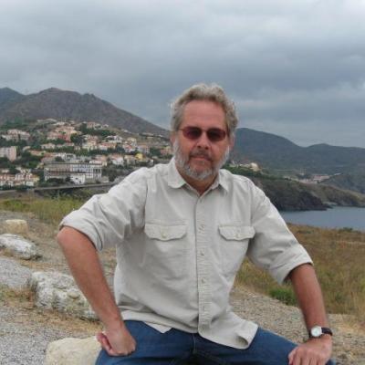 Courtesy image of Larry Ellis, sitting on a rock with a village and an ocean behind him.
